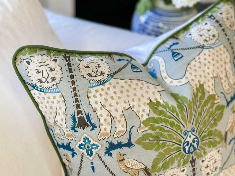May include: Decorative pillow with a light blue background and a repeating pattern of lions, palm trees, and other botanical elements. The lions are white with tan spots and blue collars. The pillow has a green border.