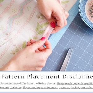 May include: A close-up of hands working with fabric and sewing supplies. The hands are holding a pink ribbon, with a pair of scissors and a blue and white bowl of pins nearby. The fabric has a floral pattern. The image includes the text "Pattern Placement Disclaimer".