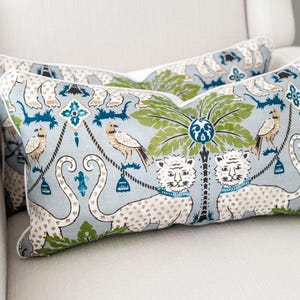 May include: Two decorative throw pillows with a light blue background and a repeating pattern of tigers, birds, and palm trees in green, blue, and white. The pillows have a white border and are placed on a white chair.