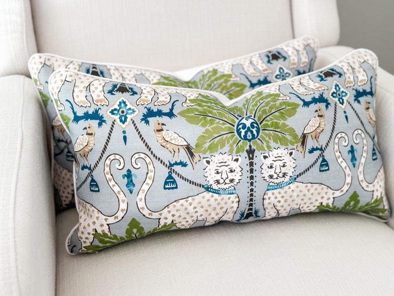 May include: Two decorative throw pillows with a light blue background and a repeating pattern of tigers, birds, and palm trees in green, blue, and white. The pillows have a white border and are placed on a white chair.
