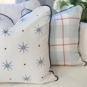 May include: Two square throw pillows with dark blue piping. The front pillow is white with a repeating pattern of blue starbursts and small red dots. The back pillow features a light blue and white plaid pattern with thin red accent lines.