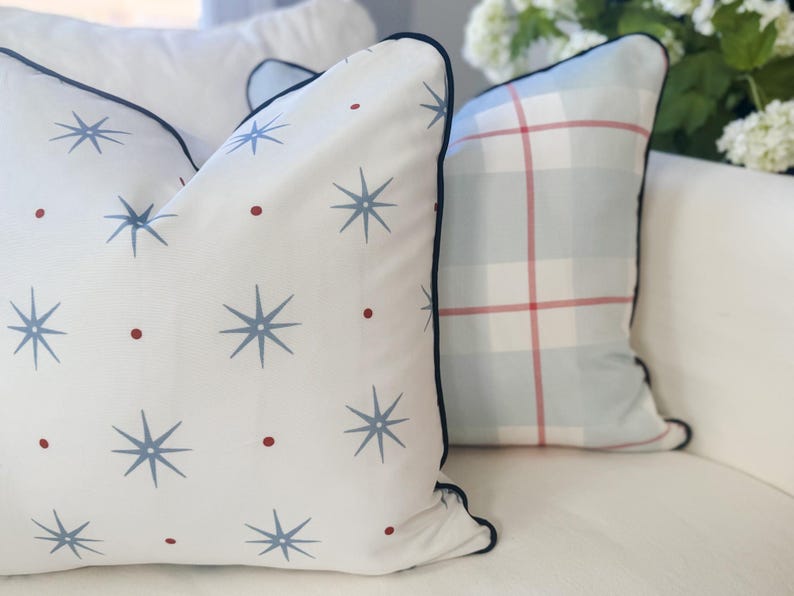 May include: Two square throw pillows with dark blue piping. The front pillow is white with a repeating pattern of blue starbursts and small red dots. The back pillow features a light blue and white plaid pattern with thin red accent lines.