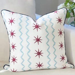 May include: A square throw pillow with a white base featuring a repeating pattern of dark red starburst shapes and vertical light blue zigzag lines. The pillow is finished with a dark blue piped edge along the perimeter of the fabric cover.