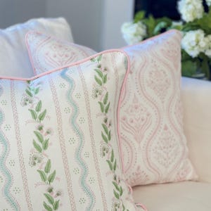 May include: Two decorative throw pillows with floral patterns. One pillow features a white background with green leaves and light blue wavy lines. The other pillow has a pink damask pattern. Both pillows have pink piping.