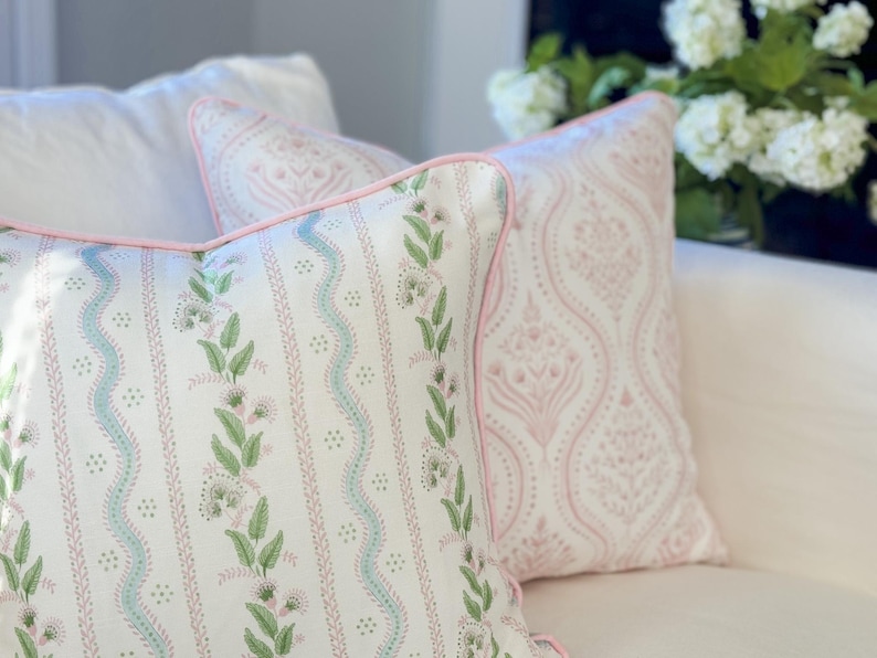 May include: Two decorative throw pillows with floral patterns. One pillow features a white background with green leaves and light blue wavy lines. The other pillow has a pink damask pattern. Both pillows have pink piping.