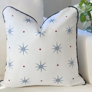 May include: A square throw pillow with a white base featuring a repeating pattern of light blue eight-pointed stars and small red dots. The pillow is finished with a dark navy blue piped edge along the perimeter, providing a clean and defined border.