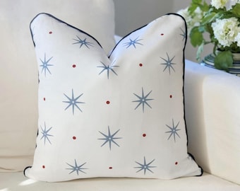 May include: A square throw pillow with a white base featuring a repeating pattern of light blue eight-pointed stars and small red dots. The pillow is finished with a dark navy blue piped edge along the perimeter, providing a clean and defined border.