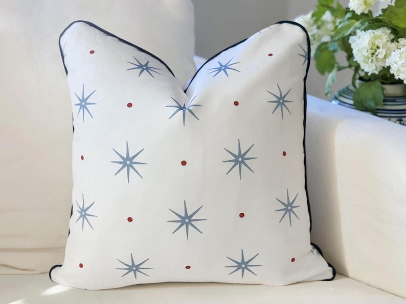 May include: A square throw pillow with a white base featuring a repeating pattern of light blue eight-pointed stars and small red dots. The pillow is finished with a dark navy blue piped edge along the perimeter, providing a clean and defined border.