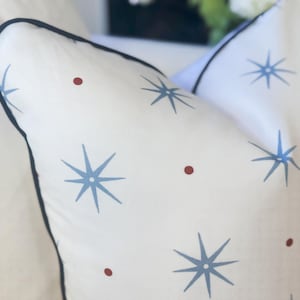 May include: A white throw pillow featuring a repeating pattern of light blue eight-pointed starbursts and small dark red circular dots. The pillow is finished with a dark navy blue piped edge that outlines the square shape of the cushion.