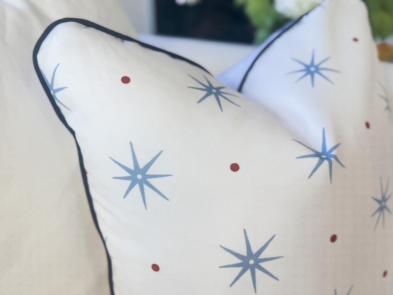 May include: A white throw pillow featuring a repeating pattern of light blue eight-pointed starbursts and small dark red circular dots. The pillow is finished with a dark navy blue piped edge that outlines the square shape of the cushion.