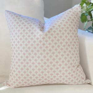 May include: A square throw pillow featuring a repeating pattern of small, dusty pink fan-shaped motifs printed on a cream-colored fabric background. The pillow has a soft, plump appearance with clean, straight edges and a consistent, symmetrical design.