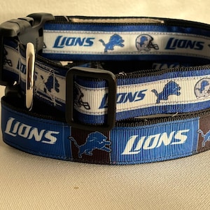 Könnte beinhalten: Zwei Hundehalsbänder übereinander gestapelt. Das obere Halsband ist schwarz mit einem blau-weißen Band mit dem Wort "Lions", einem Löwenlogo und einem Footballhelm. Das untere Halsband ist blau mit dem Wort "Lions" und einem Löwenlogo.