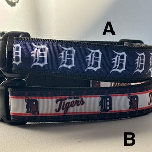 Op de afbeelding: Twee hondenriemen met zwarte plastic gespen. De bovenste halsband is marineblauw met witte "D" logo's. De onderste halsband is wit met de tekst "Detroit Tigers" en een rood en marineblauw logo.