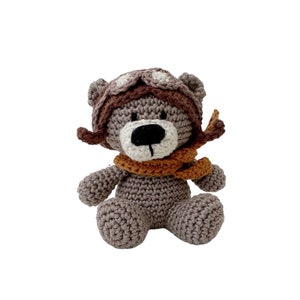 Puede incluir: Un oso de peluche gris tejido a crochet con un gorro de aviador marr&oacute;n con gafas y una bufanda marr&oacute;n y naranja.