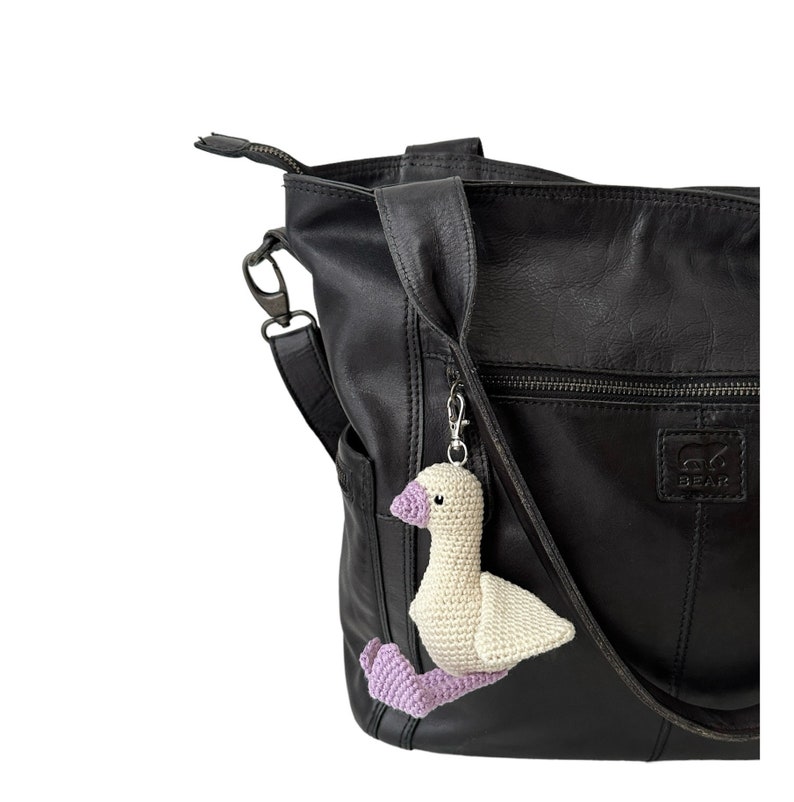 May include: A black leather tote bag with a white crocheted goose keychain attached to the strap. The bag has a zippered pocket and a logo that says "BEAR".