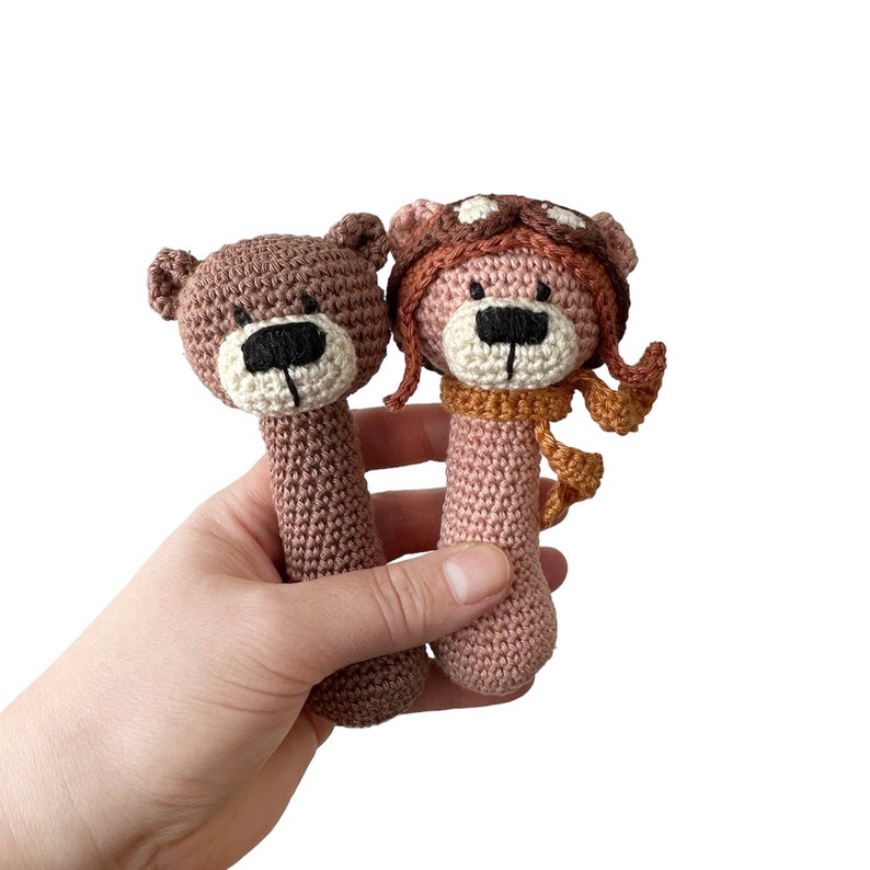 Puede incluir: Dos juguetes de sonajero de oso de crochet. Un oso es marr&oacute;n con una nariz marr&oacute;n y ojos negros. El otro oso es rosa con una nariz marr&oacute;n y ojos negros. El oso rosa lleva un gorro y una bufanda de crochet marr&oacute;n y naranja.