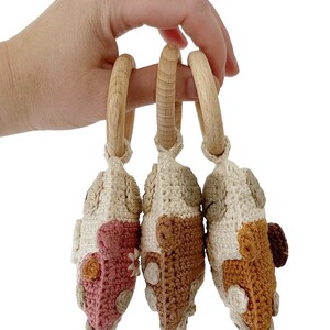 May include: Three crocheted toy cars in pink, brown, and orange with wooden rings attached. The cars are made with a cream coloured yarn and have various coloured details.