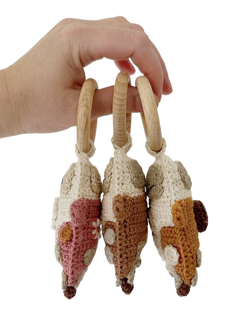 May include: Three crocheted toy cars in pink, brown, and orange with wooden rings attached. The cars are made with a cream coloured yarn and have various coloured details.
