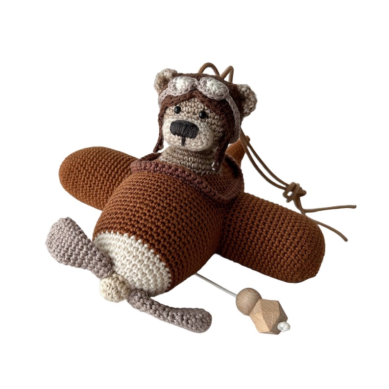 Puede incluir: Un juguete de avi&oacute;n de crochet marr&oacute;n con un oso marr&oacute;n que lleva un gorro de aviador marr&oacute;n y gafas de protecci&oacute;n en la cabina. El avi&oacute;n tiene una h&eacute;lice beige y una varilla de madera.