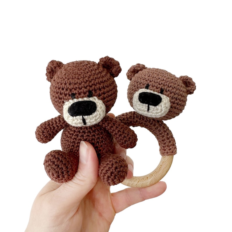 Puede incluir: Un sonajero de oso de crochet marr&oacute;n con un anillo de madera. El oso tiene ojos negros y una nariz blanca.