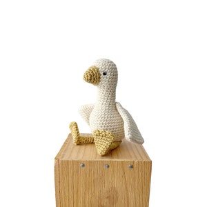 May include: A white crocheted goose with yellow feet sitting on a wooden box.