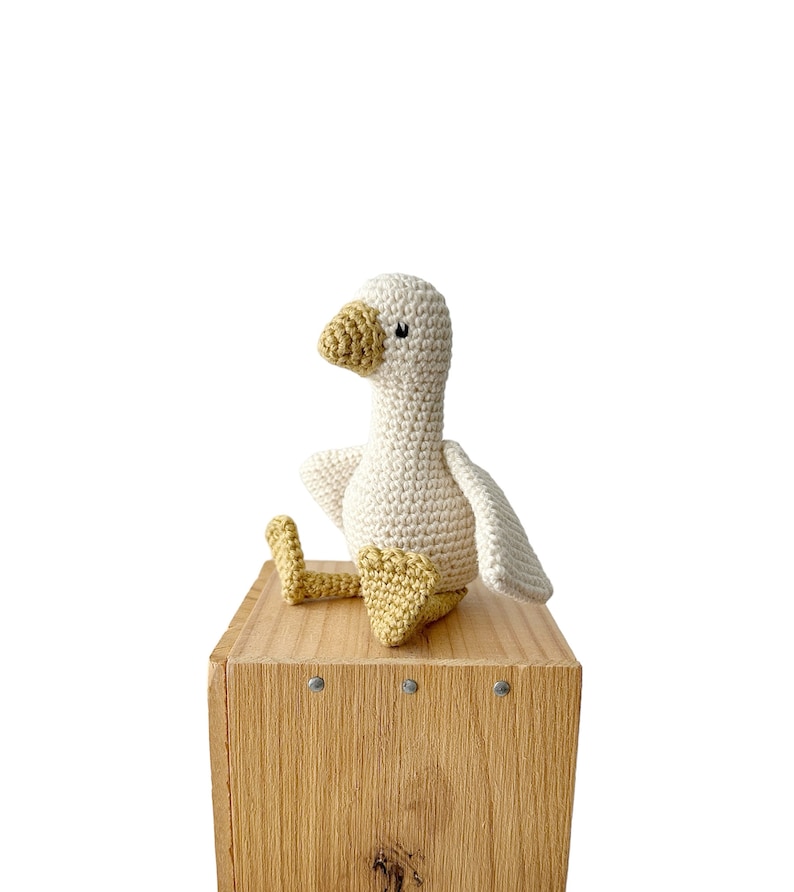 May include: A white crocheted goose with yellow feet sitting on a wooden box.