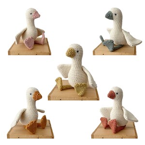 May include: Five crocheted goose toys in different colors, each sitting on a wooden shelf. The geese have different colored beaks and feet.