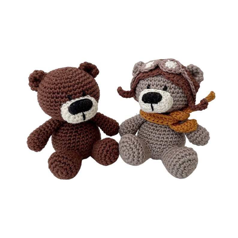 Puede incluir: Dos osos de peluche tejidos a crochet, uno marr&oacute;n y otro gris. El oso gris lleva un gorro de aviador marr&oacute;n con detalles blancos y una bufanda naranja.