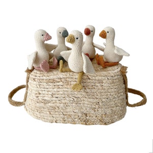 May include: Five crocheted white geese with colorful accents sitting in a woven basket. The geese have different colored beaks and feet.