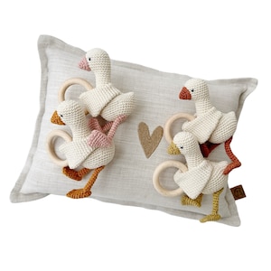 May include: Four crocheted goose rattle toys with wooden rings. The geese are white with pink, orange, and yellow accents. The toys are arranged on a beige pillow with a brown heart.
