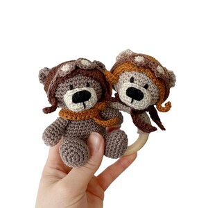 Puede incluir: Dos osos de peluche de crochet marrones que llevan gorros de aviador y bufandas est&aacute;n unidos a un anillo de madera. Los osos se miran y tienen ojos y narices negros.