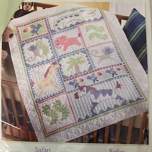 May include: A Bucilla Safari stamped cross stitch crib cover kit featuring a white quilted fabric with a grid of animal illustrations including a crocodile, elephant, monkeys, giraffe, and toucans. The packaging states dimensions of 34 inches by 43 inches.