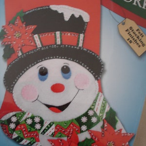 May include: A felt stocking craft kit featuring a snowman with a red nose and green and white scarf. The stocking is 18 inches long and includes felt, sequins, cotton floss, needles, and patterns. The kit is titled "Felt Stocking Finishes 18" and includes the word "Noel" at the top.