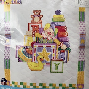 May include: A baby quilt kit featuring a colorful illustration of a clown surrounded by toys, including a teddy bear, a duck, and blocks that spell out "BABY". The kit includes pre-bound poly/cotton fabric, embroidery floss, a needle, and complete instructions. The kit is 34 inches by 43 inches.