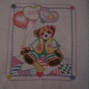May include: A cross-stitch pattern for a patchwork bear quilt. The design features a brown bear holding three balloons with hearts and the word "XOX" on them. The bear is wearing a green vest with pink and yellow flowers. The quilt is made up of different colored squares with a white background.