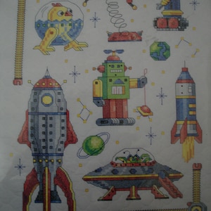 May include: A colorful cross-stitch pattern featuring a variety of space-themed characters, including a rocket, a UFO, a robot, and an alien. The pattern is set against a white background with yellow stars.
