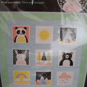 May include: A set of nine cross-stitch quilt blocks featuring nature-themed designs. The blocks include a panda, sun, teepee, bunny, trees, bear, rainbow, mountains, and a cloud with sprinkles. The text "First you stitch. Then you snuggle." is printed on the packaging.