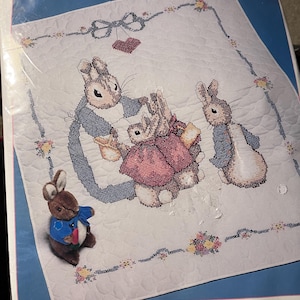 May include: A cross stitch kit featuring Mrs. Rabbit and Bunnies from Beatrix Potter. The kit includes a printed cross stitch design on a 35 by 43 inch quilt, 100% cotton floss, a needle, and complete instructions.