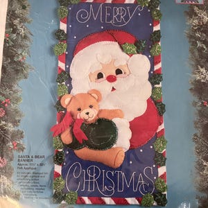 May include: A felt appliqué Christmas banner kit featuring Santa Claus holding a teddy bear. The banner has a blue background with the words "MERRY CHRISTMAS" and measures approximately 11 inches by 22 inches.