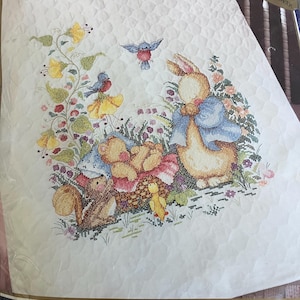 May include: A Bucilla Baby stamped cross stitch kit for a 34 inch by 43 inch crib cover. The white quilted fabric features a printed design of a rabbit in a blue jacket, a teddy bear, a squirrel, a duckling, and two blue birds among flowers.
