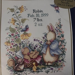 May include: A Bucilla Baby counted cross stitch kit featuring a birth record design with a rabbit, a teddy bear in a basket, a squirrel, and a small bird. The text reads Robin, Feb. 18, 1999, 7 lbs. 2 oz. surrounded by colorful floral illustrations.