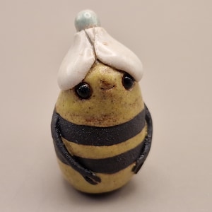 May include: A small, pear-shaped ceramic figurine resembling a bee with a yellow body, two horizontal black stripes, and two black arms. It features two dark, glossy eyes, a tiny mouth, and a white, flower-like hat topped with a small blue sphere.