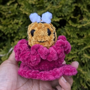 May include: A handmade crochet bee with a yellow and black body, white antennae, and a magenta flower-like base. The plush toy is held in a hand against a backdrop of green foliage.