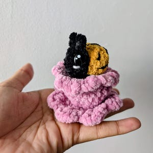 May include: A handmade plush toy featuring a black bunny and a yellow jack-o'-lantern face, nestled in a pink, ruffled, plush container. The bunny has large white eyes. The toy is held in a hand.