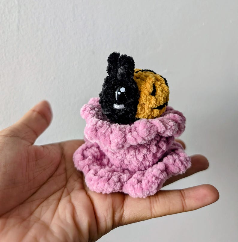 May include: A handmade plush toy featuring a black bunny and a yellow jack-o'-lantern face, nestled in a pink, ruffled, plush container. The bunny has large white eyes. The toy is held in a hand.