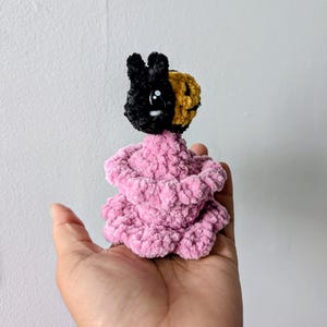 May include: A handmade crochet doll with a black and gold head and a pink body. The doll has a bunny-like appearance with a smiling face. The doll is made of soft, textured yarn.