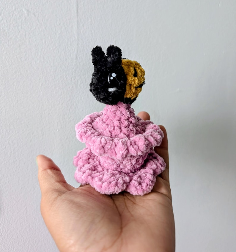 May include: A handmade crochet doll with a black and gold head and a pink body. The doll has a bunny-like appearance with a smiling face. The doll is made of soft, textured yarn.