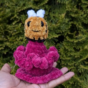 May include: A handmade plush bee toy with a fuzzy, textured appearance. The bee features a yellow and black head with white antennae and a magenta body. The toy is held in a hand against a backdrop of green foliage.
