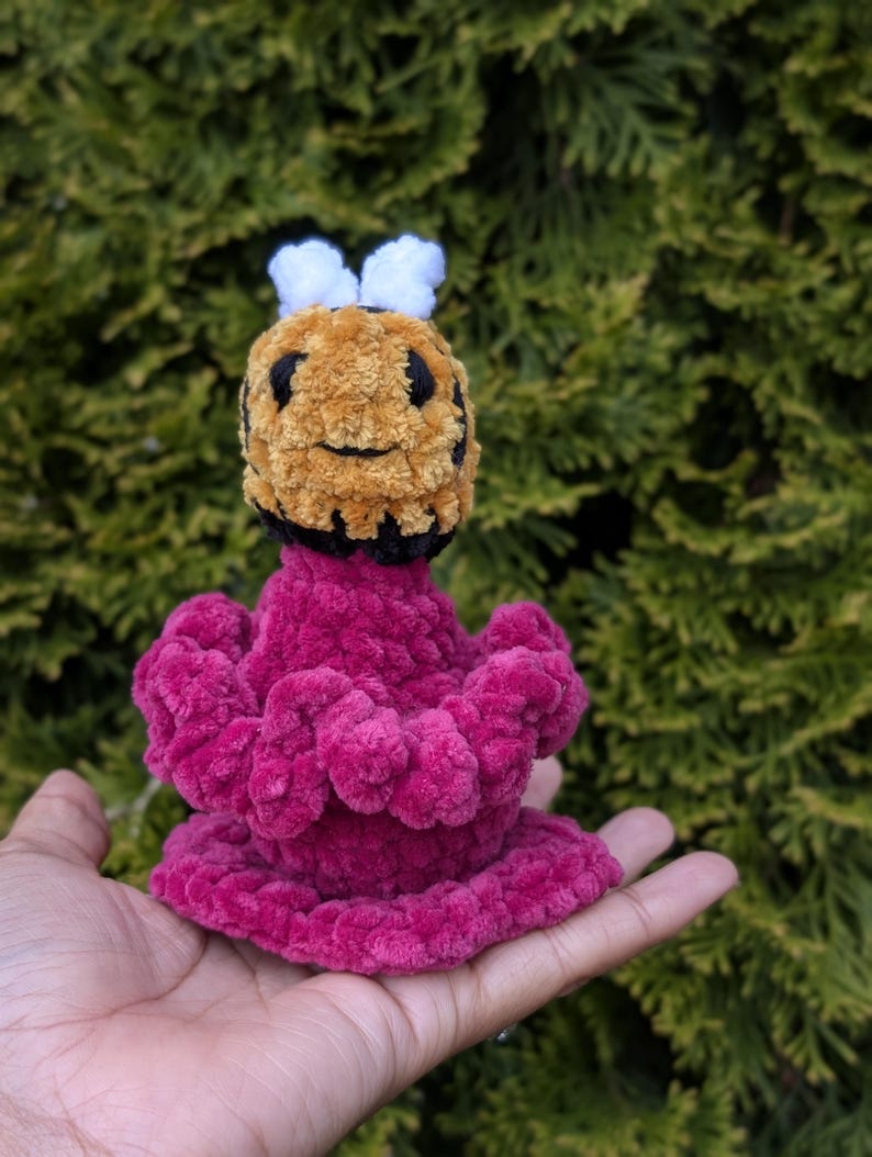 May include: A handmade plush bee toy with a fuzzy, textured appearance. The bee features a yellow and black head with white antennae and a magenta body. The toy is held in a hand against a backdrop of green foliage.