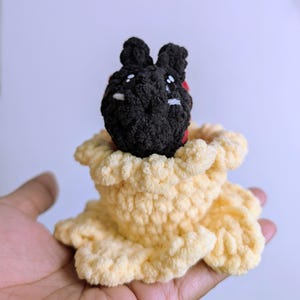 May include: A handmade crochet bunny with a black head and white eyes, nestled in a light yellow crochet flower. The bunny has red accents and is made of soft yarn.